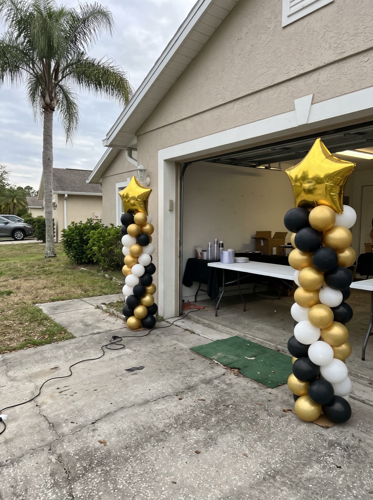 Two spiral balloon columns in black and gold with star toppers beside a garage door at a grad party
